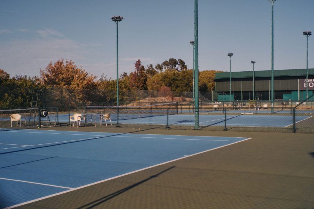 Multiple outdoor tennis courts with blue playing surfaces, nets, surrounding fences, and tall lighting poles are visible.